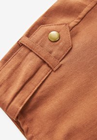 Tissu couleur rouille avec un onglet cousu fixé par un bouton-pression rond en laiton, montrant une texture détaillée et des lignes de couture.