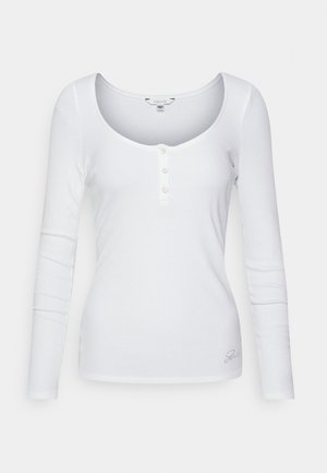 Chemise Henley blanche à manches longues côtelée avec encolure dégagée, trois boutons sur le devant et petit logo près de l'ourlet inférieur.