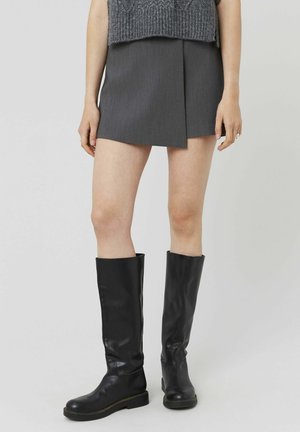 Jupe mini grise avec un pli diagonal à l'avant, associée à des bottes noires en cuir hautes avec semelle plate et design simple.