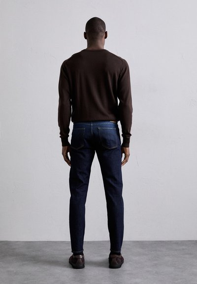 Suéter marrón, jeans azul oscuro, corte ajustado. Los jeans tienen bolsillos traseros con detalle de costura, manos relajadas a los lados, fondo minimalista.