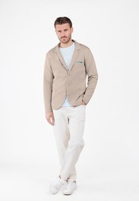 Mann mit übereinandergeschlagenen Beinen, der ein beiges Blazer, ein hellblaues T-Shirt, weiße Hosen und weiße Sneaker vor einem schlichten weißen Hintergrund trägt.