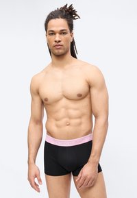 Φορώντας μαύρα μποξέρ με ροζ ζώνη μέσης, με την επωνυμία "LACOSTE", ένας γυμνασμένος άνδρας με κοτσίδες στέκεται μπροστά σε απλό φωτεινό φόντο.