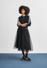 Black knitted sweater with white stripes, black tulle skirt, black socks with white stripes, and black shoes with gum sole, standing on blue carpet.