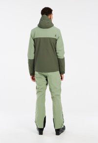 Grünes zweiteiliges Oberbekleidungsset mit einer Kapuzenjacke, die einen kontrastierenden dunkelgrünen oberen Teil und passende Hosen aufweist, gefertigt aus glattem, strapazierfähigem Material.