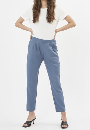 Femme portant un haut blanc à manches courtes, un pantalon plissé bleu taille haute et des sandales noires à talons, debout les mains sur les hanches.