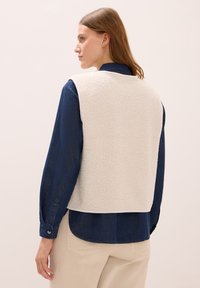 Fleece svetr v krémové barvě, s měkkou texturou a bez rukávů, nošený přes tmavě modrou džínovou košili s knoflíkovými manžetami a přímým spodním lemem.