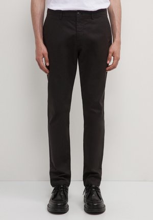Schwarze, schlanke Hose mit glatter Textur, ausgestattet mit einem Knopfverschluss und Gürtelschlaufen. Kombiniert mit schwarzen Schnürschuhen.