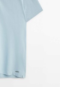 Lichtblauw polohemd met korte mouwen, textuurrijke stof, rechte zoom en een klein metalen merkembleem vlak bij de onderkant.