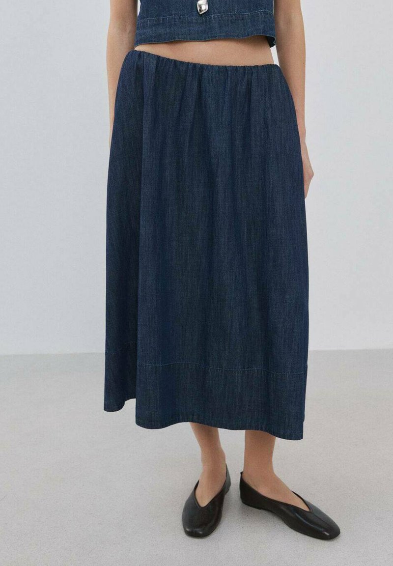 Trapera midi su elastičnim pojasom, tamnoplava boja, A-oblika, s kontrastnim šivanjem. Uparena s crnim baletankama.