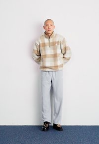 Fleece pulover v bež in rjavem karirastem vzorcu s pol-zadrgo; v kombinaciji s svetlo sivimi trenirkami in črnimi čevlji z vzorcem.