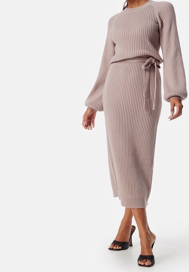 Femme portant un top beige en maille côtelée à manches longues et une jupe midi assortie avec un lien sur le côté, associée à des sandales à talons noires ouvertes à l’avant.