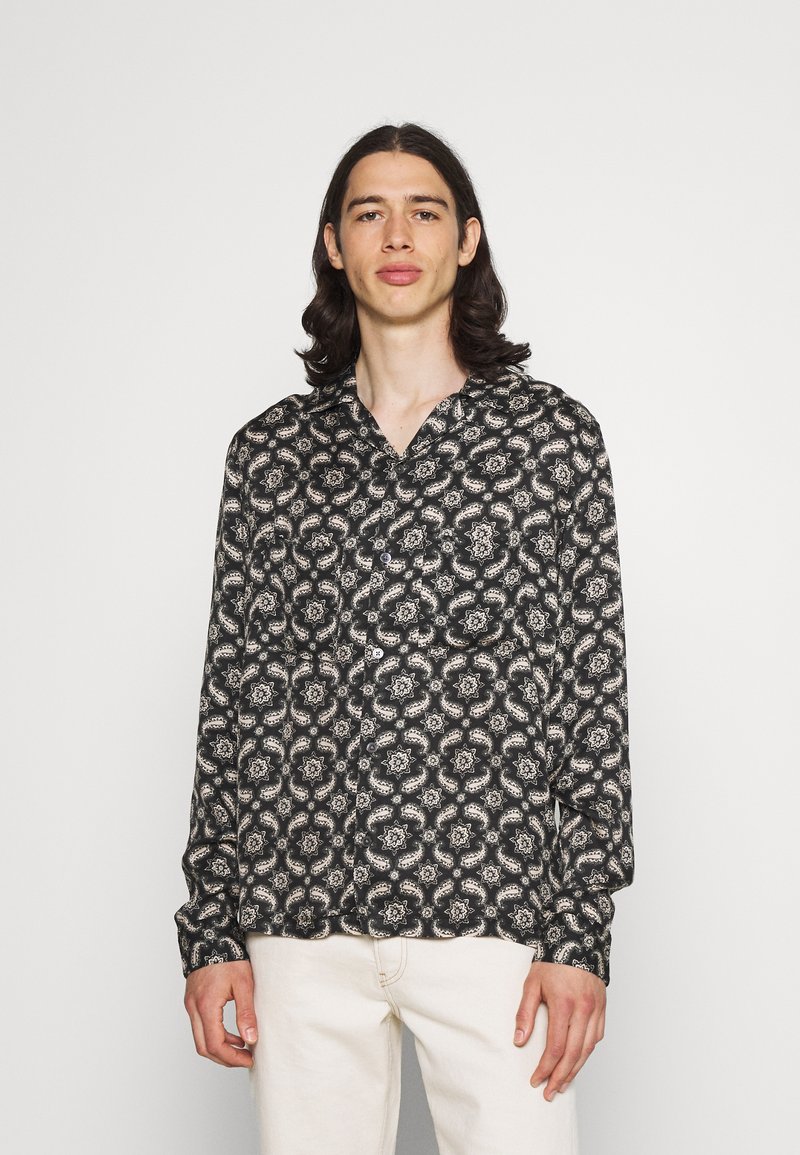 Chemise à manches longues en tissu noir avec un motif floral blanc, dotée d'un col et de détails de boutons. Portée avec un pantalon clair.