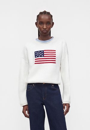 Γυναίκα με πλεξούδες, φορεί ένα λευκό πουλόβερ με τη σημαία των Ηνωμένων Πολιτειών και σκούρα μπλε τζιν, στέκεται μπροστά σε απλό γκρι φόντο.