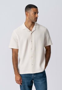 Kortærmet cremet skjorte med knaplukning og krave, der har ribbet tekstur og en lidt afslappet pasform, parret med blå jeans.