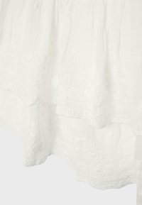 Tissu blanc avec motifs floraux brodés et bords festonnés, montrant des textures stratifiées et des détails de couture délicats.