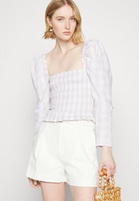 Blusa xadrez lilás claro com mangas bufantes e corpete franzido, combinada com calções brancos plissados. Acessórios incluem um saco de contas de madeira.