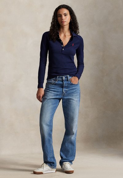 Tamsiai mėlynas ilgomis rankovėmis polo marškinėliai su sagų eilute, derinami su šviesiai mėlynais tiesiais džinsais ir baltais sportiniais bateliais, nufotografuota neutraliame fone.