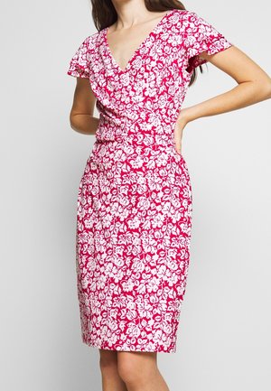 Frau trägt knie- langes pinkes Kleid mit weißem Blumenmuster, V-Ausschnitt und kurzen flatternden Ärmeln, steht mit einer Hand in der Hüfte.