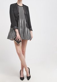 Femme portant une robe à carreaux noir et blanc avec un blazer noir, tenant une pochette noire et dorée, et des talons hauts noirs.