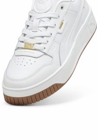 Baskets blanches avec tige en cuir lisse, écusson rond avec logo, accent doré sur les lacets et semelle en caoutchouc marron à texture rainurée.