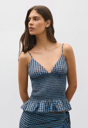 Giovane donna con lunghi capelli scuri che indossa un top smanicato a quadri blu e marroni e una gonna abbinata su sfondo chiaro e semplice.