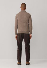 Kabeltrui in taupe met hoge kraag, gedragen met donkere broeken en bruine schoenen. Geweven structuur zichtbaar in het breipatroon.