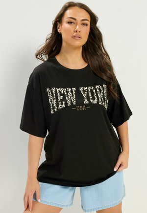 Kvinde med langt brunt hår iført sort oversized T-shirt med leopardprint "New York USA" tekst og lyseblå cowboyshorts.