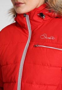Chaqueta acolchada roja con capucha forrada de piel, cremalleras plateadas y detalle de logo en el pecho. El material parece elegante y resistente a la intemperie.