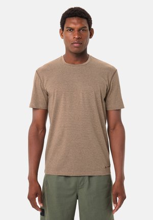 Ein Mann, der nach vorne schaut, trägt ein hellbraunes, gestreiftes Kurzarmhemd und olivgrüne Hosen vor einem schlichten weißen Hintergrund.