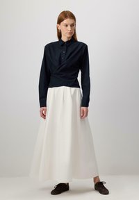 Blusa a maniche lunghe di colore blu navy con bottoni e design avvolgente, abbinata a una gonna bianca fluente. Scarpe marroni completano l'outfit. Semplice ed elegante.