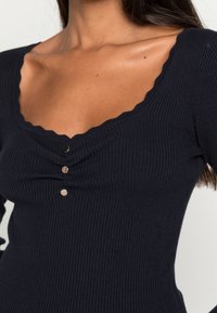Haut côtelé à manches longues bleu marine avec un décolleté festonné, orné de trois boutons métalliques au centre. Texture douce et coupe ajustée.