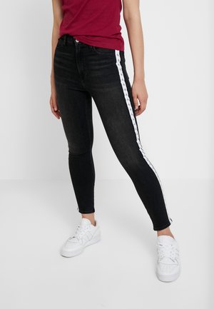 Osoba nosząca czarne obcisłe dżinsy z białym paskiem z logo po boku, w połączeniu z białymi trampkami i czerwonym topem, stojąca na jednolitym tle.