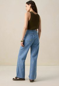 Femme portant un top sans manches en ribbed vert, un jean large bleu clair avec des taches brodées jaunes, des sandales et des boucles d'oreilles en or, se tenant de profil.