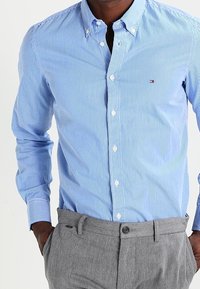 Camisa de botón a rayas azul claro con cuello y botones blancos. Combinada con pantalones grises, que presentan un diseño sencillo y una tela texturizada.