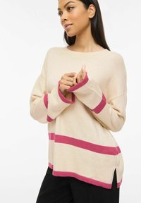 Femme portant un pull beige avec des rayures roses sur les manches, l'ourlet et le milieu du vêtement, associée à un pantalon noir, posant les mains près de la poitrine.