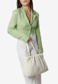 Witte textuur tote bag met golvend ontwerp, gecombineerd met een lichtgroene cropped blazer en witte broek, vastgehouden in de hand.