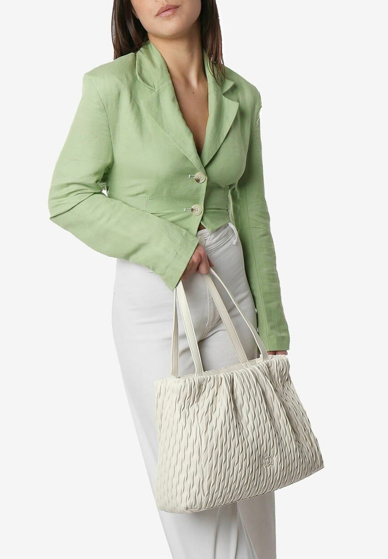Witte textuur tote bag met golvend ontwerp, gecombineerd met een lichtgroene cropped blazer en witte broek, vastgehouden in de hand.
