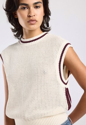 Jong persoon die een mouwloze crèmekleurige gebreide trui draagt met bordeauxrode strepen op de kraag en armsgaten, staand tegen een effen achtergrond.