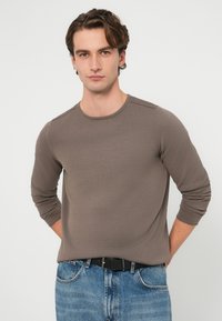 Koszulka z długim rękawem i okrągłym dekoltem w jasnobrązowym kolorze, wykonana z teksturowanej tkaniny. Połączona z jasnoniebieskimi dżinsami i czarnym paskiem.