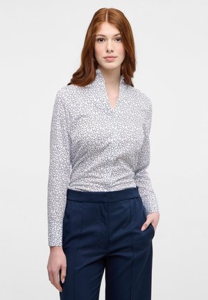 Camicia bianca a maniche lunghe con motivo a spirale nera; presenta un colletto scialle; abbinata a pantaloni su misura blu navy, il tessuto appare liscio.