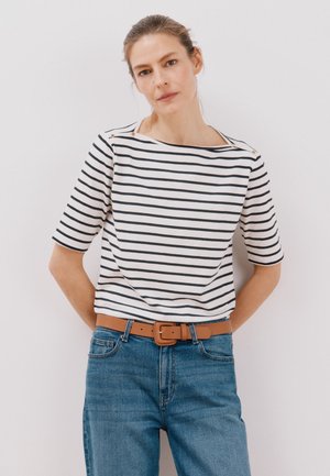 T-shirt en coton rayé présentant des rayures horizontales noires et blanches, un col bateau et des accents dorés aux épaules. Porté avec un jean bleu et une ceinture beige.