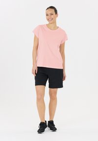 Lichtroze, T-shirt met korte mouwen gecombineerd met zwarte shorts en zwarte sportieve schoenen, op een model tegen een effen witte achtergrond.