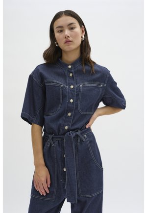 Denim-Overall mit kurzen Ärmeln, dunkelblauer Farbe, Vorderknöpfen, zwei Brusttaschen und einem gebundenen Taillengürtel. Kontrastnähte.