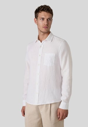 Mann trägt ein weißes Hemd mit langen Ärmeln und Brusttasche, kombiniert mit beigen Hosen, vor einem schlichten grauen Hintergrund.