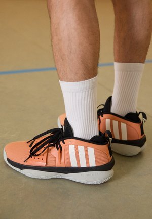 Chaussures de sport adaptatives en pêche, avec dessus en mesh, accents noirs, trois rayures blanches et semelle en caoutchouc. Portées avec des chaussettes blanches mi-mollet.