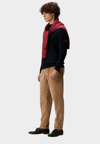 Marineblauer Strickpullover, roter Schal über die Schultern gelegt, beiger Cordhosen, schwarze Schuhe, Modell steht seitlich vor einem grauen Hintergrund.