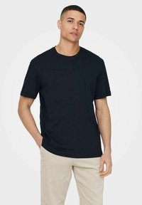 Schwarzes Baumwoll-T-Shirt mit rundem Halsausschnitt und kurzen Ärmeln, kombiniert mit hellbeigen Cordhosen. Minimalistisches Design, keine sichtbaren Muster.