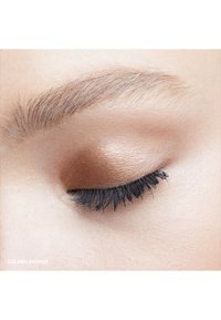Nahaufnahme eines Auges mit schimmerndem goldbraunem Lidschatten, schwarzem Mascara auf langen Wimpern und natürlichen Brauenhaaren darüber.