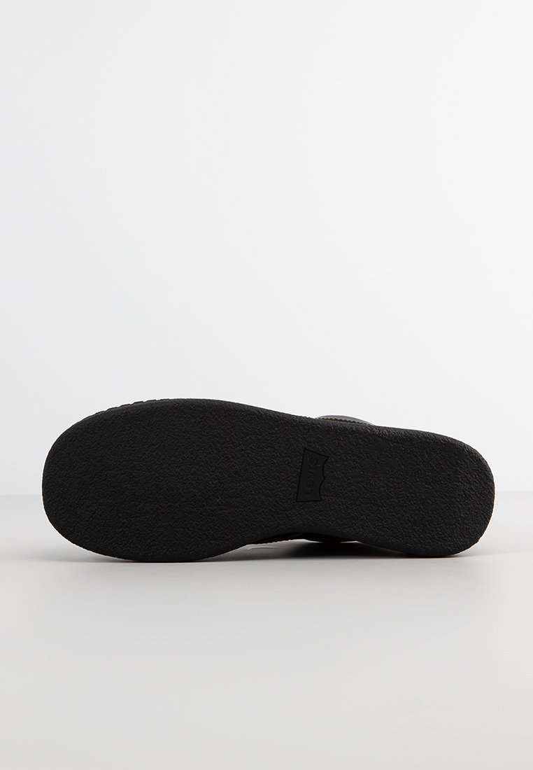 Semelle de chaussure en caoutchouc noir avec une surface texturée pour une meilleure adhérence, portant une empreinte de logo. La forme est plate et arrondie au bout.