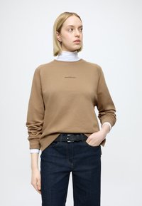 Καφέ βαμβακερό φούτερ με στρογγυλή λαιμόκοψη και μακριά μανίκια, με μικρό λογότυπο στο στήθος, συνδυασμένο με σκούρο τζιν και μαύρη ζώνη.